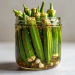 Close-up of a clear glass jar filled with vibrant green pickled okra pods, submerged in brine with visible spices and garlic cloves.