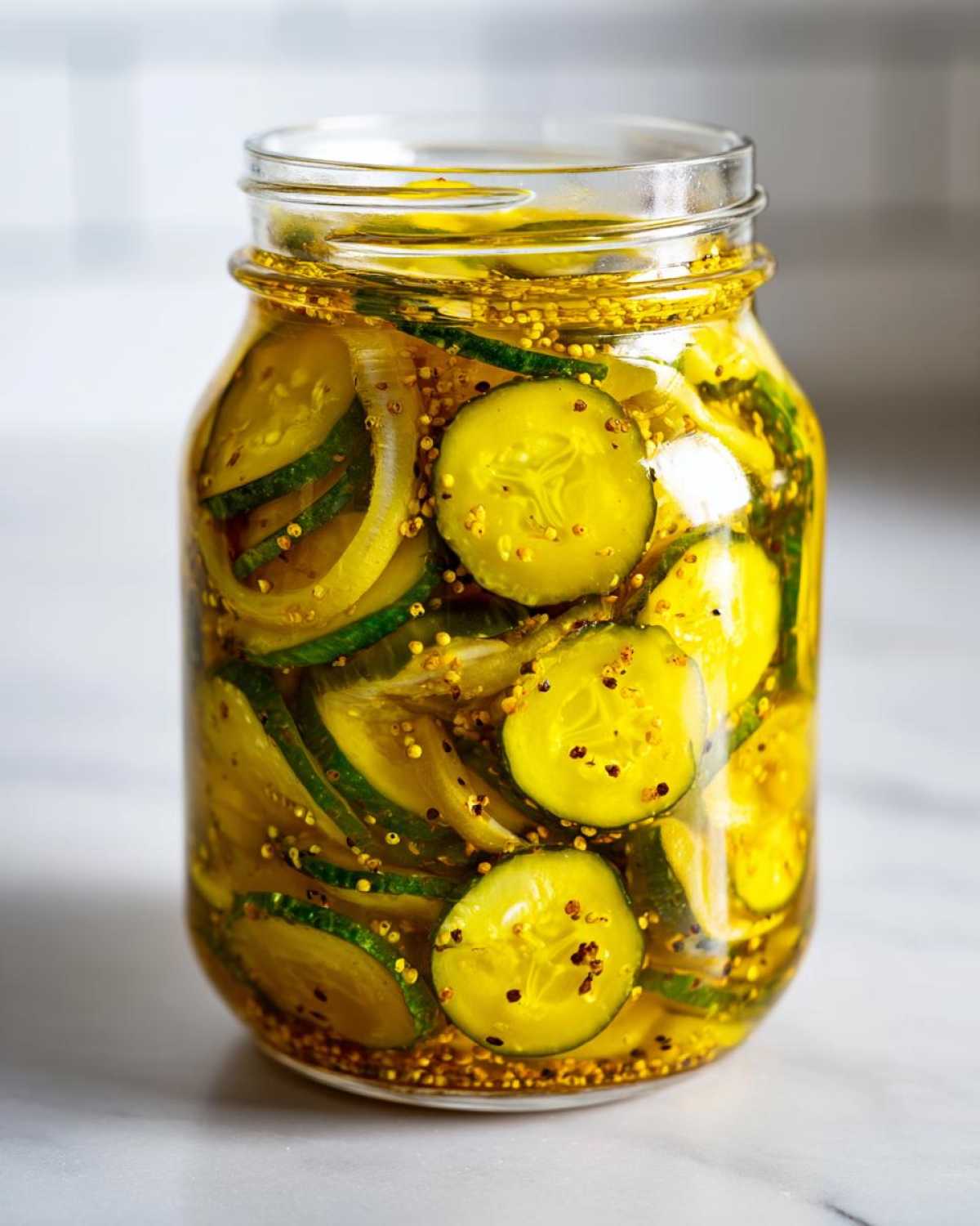 A clear glass jar filled with sliced bread and butter pickles, onions, and mustard seeds in a golden brine.