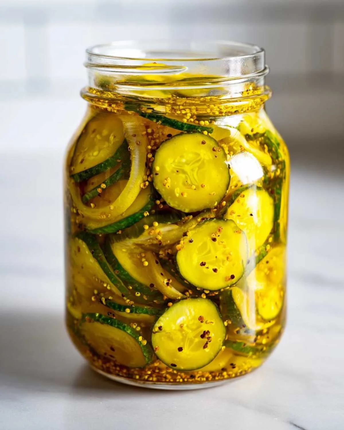 A clear glass jar filled with sliced bread and butter pickles, onions, and mustard seeds in a golden brine.