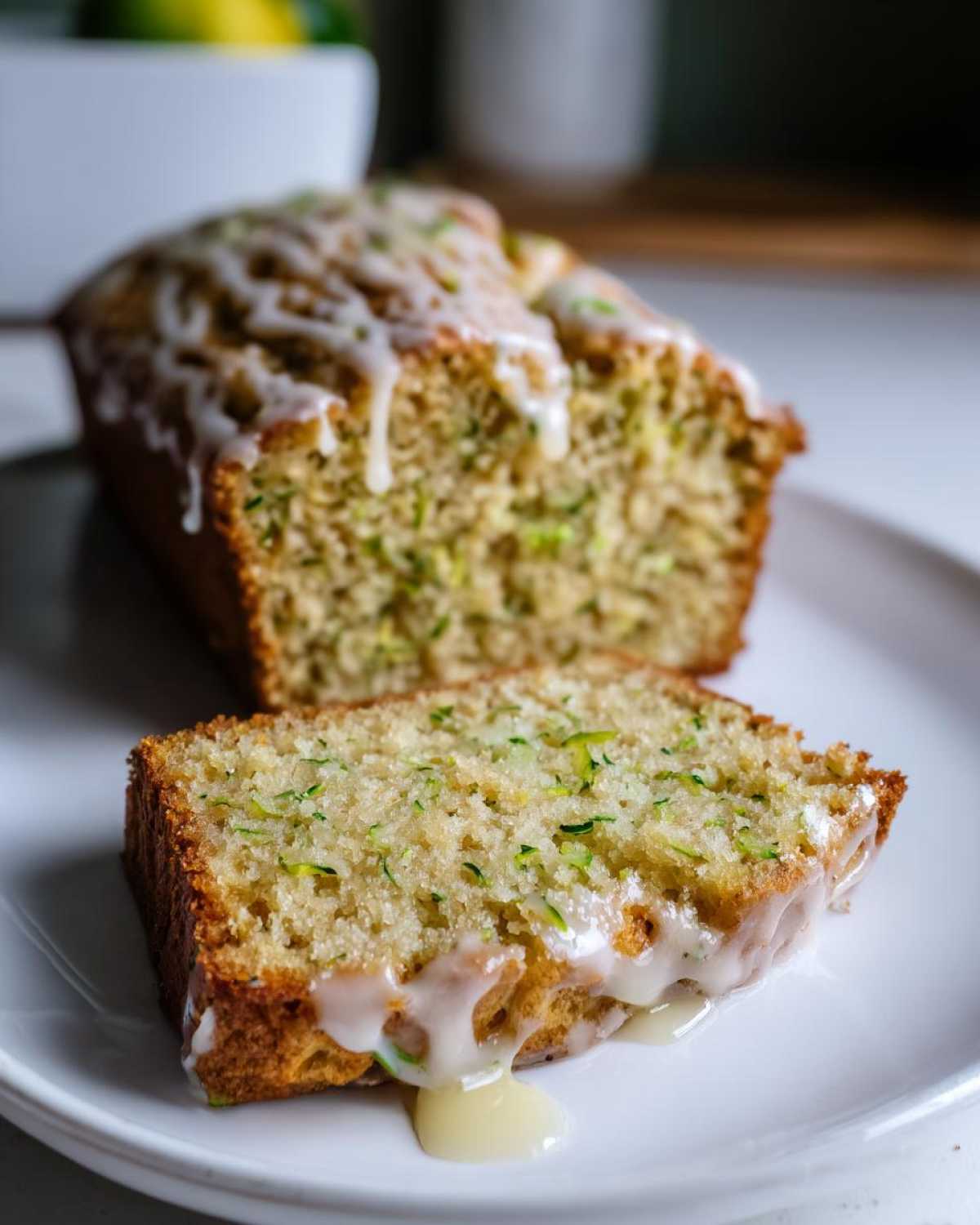 A whole loaf of lemon zucchini bread with glaze and a slice of lemon zucchini bread on a white plate.