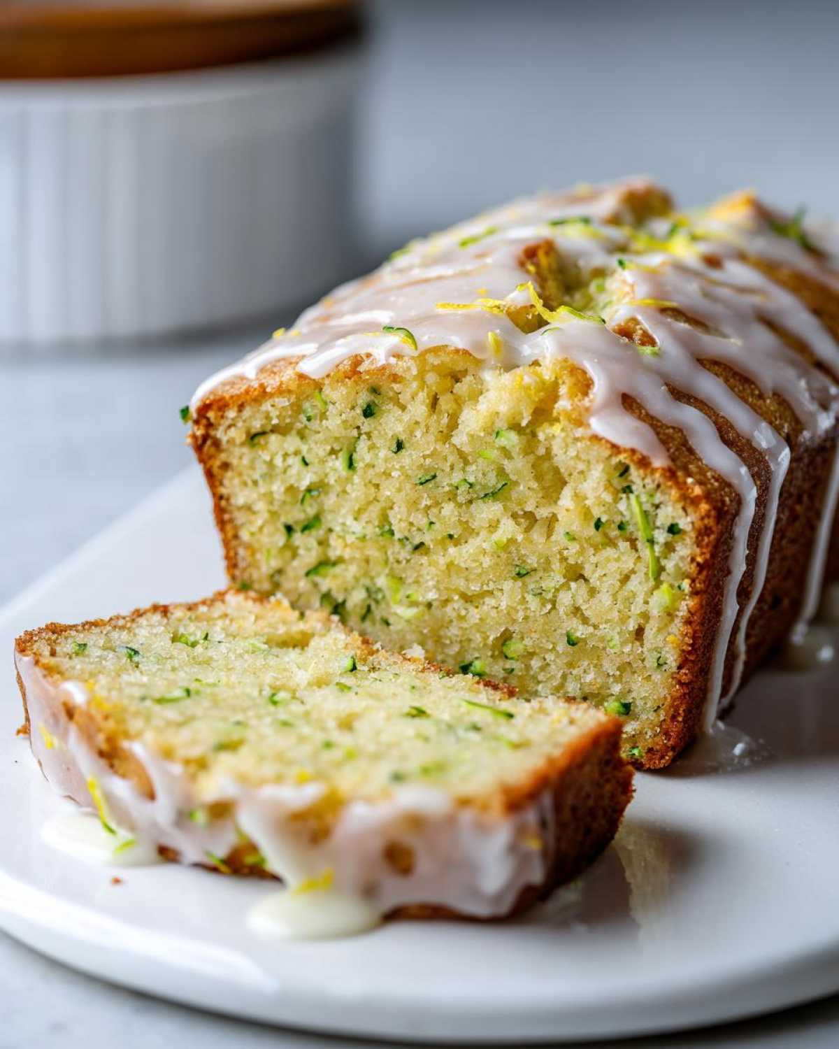 A close-up of a whole loaf of lemon zucchini bread with a slice cut, both drizzled with white glaze and lemon zest.