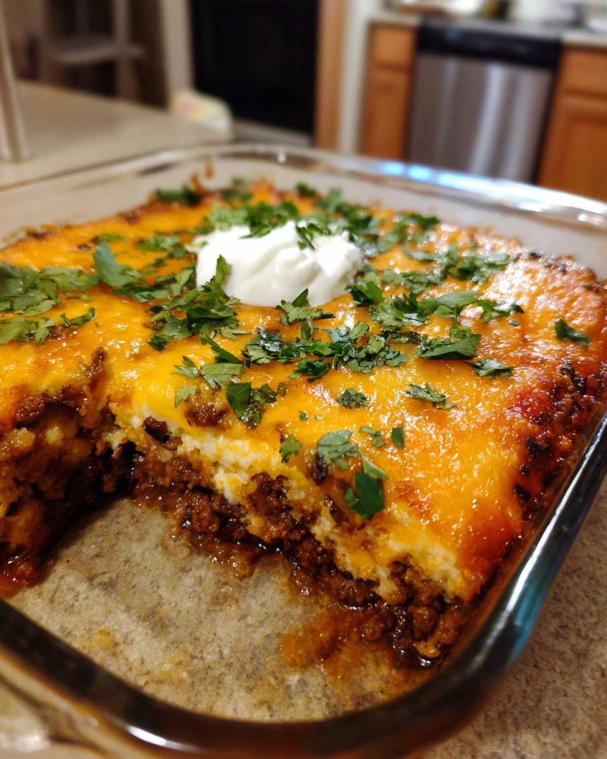 Close-up of a Mexican casserole in a glass baking dish, with a scoop removed showing layers of meat, cheese, and a dollop of sour cream and fresh cilantro on top.