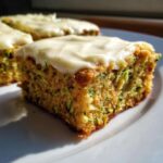 A close-up shot of a single square zucchini bar on a white plate, topped with creamy white frosting. Visible specks of green zucchini are throughout the moist cake.