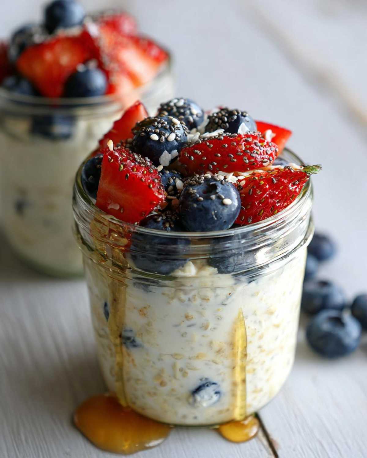 A glass jar filled with overnight oats, topped with fresh strawberries, blueberries, and chia seeds, with honey drizzled down the side.