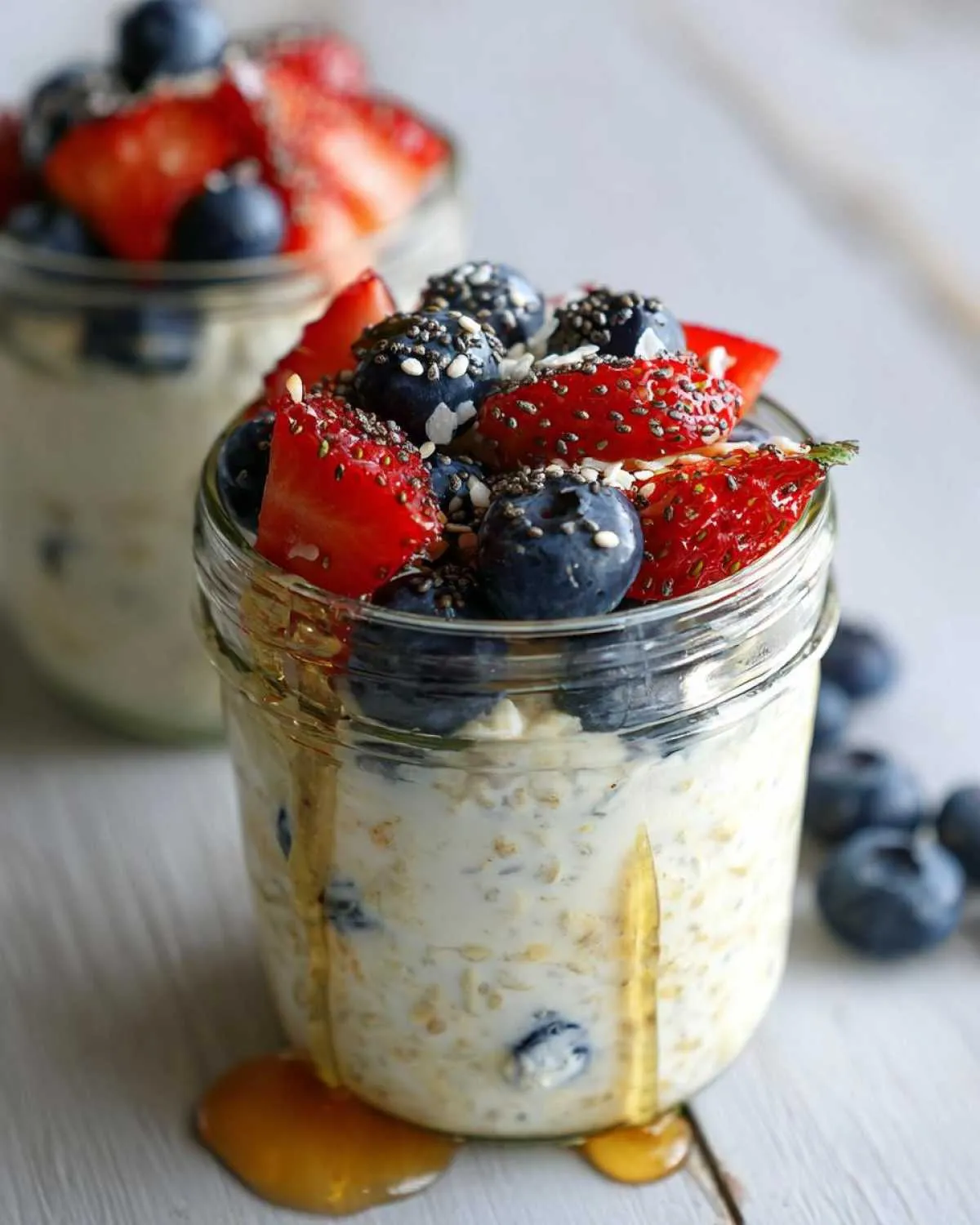 A glass jar filled with overnight oats, topped with fresh strawberries, blueberries, and chia seeds, with honey drizzled down the side.