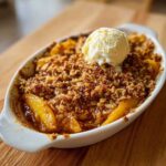 A close-up shot of a golden-brown peach crisp in a white baking dish, topped with a scoop of melting vanilla ice cream.