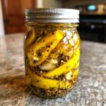 A clear glass jar filled with bright yellow pickled banana peppers, garlic cloves, and various pickling spices in a brine.