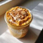 Close-up of a glass jar filled with creamy Pumpkin overnight oats, topped with white cream and a sprinkle of cinnamon.