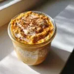 Close-up of a glass jar filled with creamy Pumpkin overnight oats, topped with white cream and a sprinkle of cinnamon.