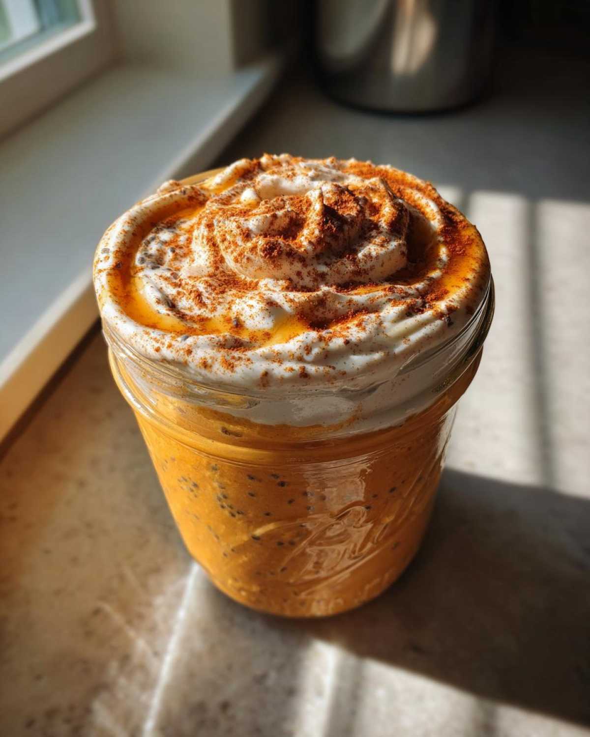 A close-up of a glass jar filled with creamy pumpkin overnight oats, topped with whipped cream, cinnamon, and a drizzle of syrup.