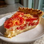 A close-up of a single slice of tomato pie on a white plate, showing layers of red tomatoes and creamy filling.