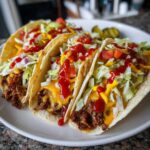Three smash burger tacos on a white plate, topped with cheese, lettuce, tomato, ketchup, and mustard.