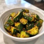 A white bowl filled with vibrant green pieces of smashed cucumber salad, coated in a reddish-brown sauce and sprinkled with white sesame seeds.