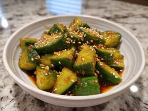 Close-up of a white bowl filled with vibrant green smashed cucumber salad, coated in a reddish-brown sauce and sprinkled with white sesame seeds.