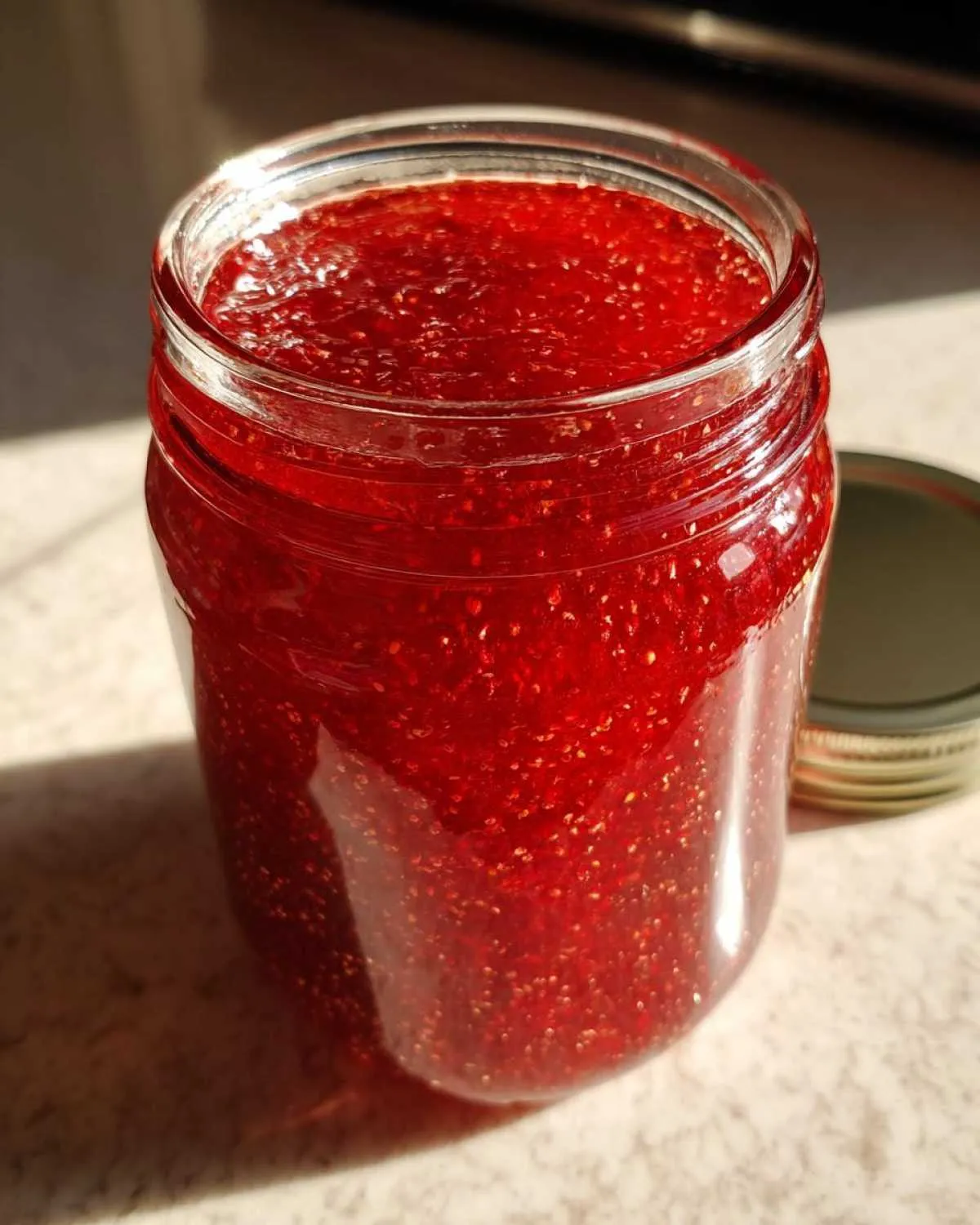 Close-up view of a glass jar filled with bright red strawberry jam, with the lid off to the side.