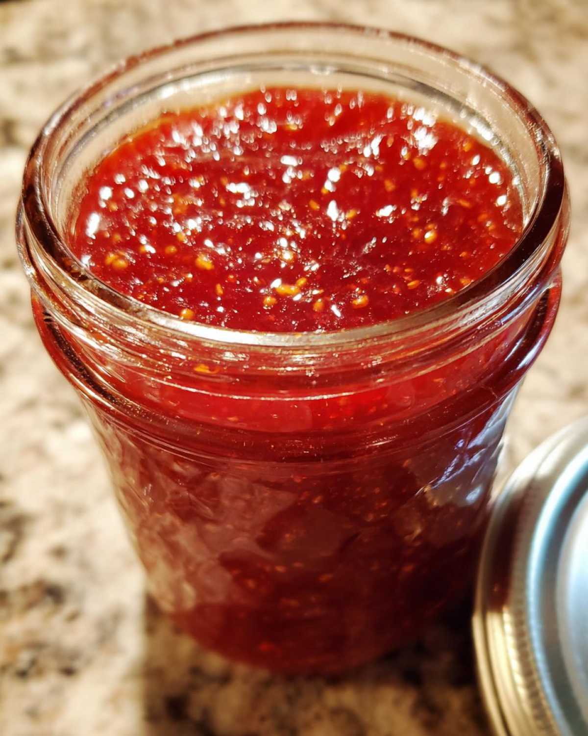 Close-up of a glass jar filled with bright red strawberry jam, showing seeds and texture.