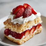 A close-up of a single slice of strawberry shortcake with layers of golden cake, fresh sliced strawberries, and whipped cream.
