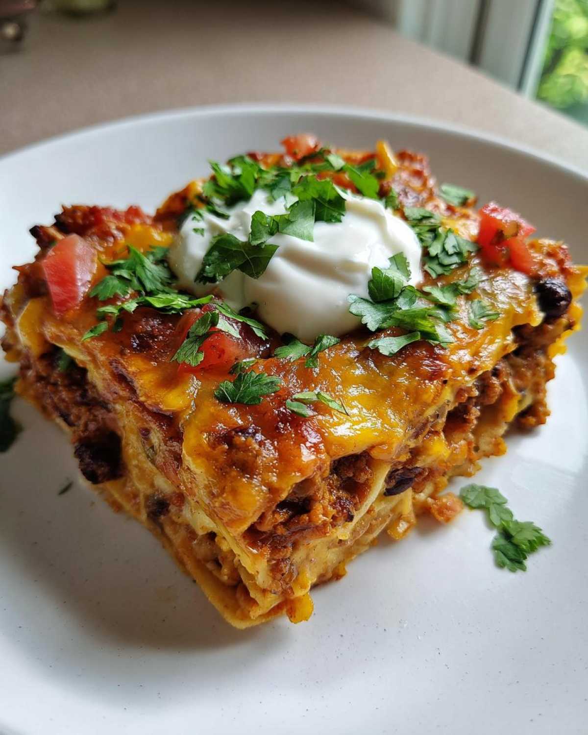 Close-up of a slice of taco casserole topped with sour cream, diced tomatoes, and fresh cilantro.