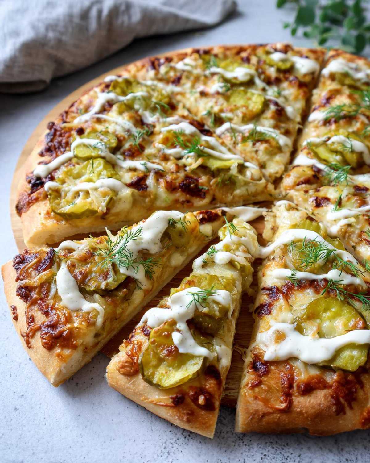 Close-up view of sliced pickle pizza topped with cheese, pickles, dill, and white sauce.