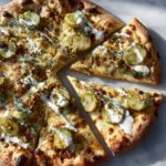 Overhead shot of a whole pickle pizza with several slices cut, topped with dill, sliced pickles, and a creamy white sauce.