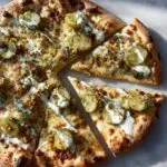 Overhead shot of a whole pickle pizza with several slices cut, topped with dill, sliced pickles, and a creamy white sauce.