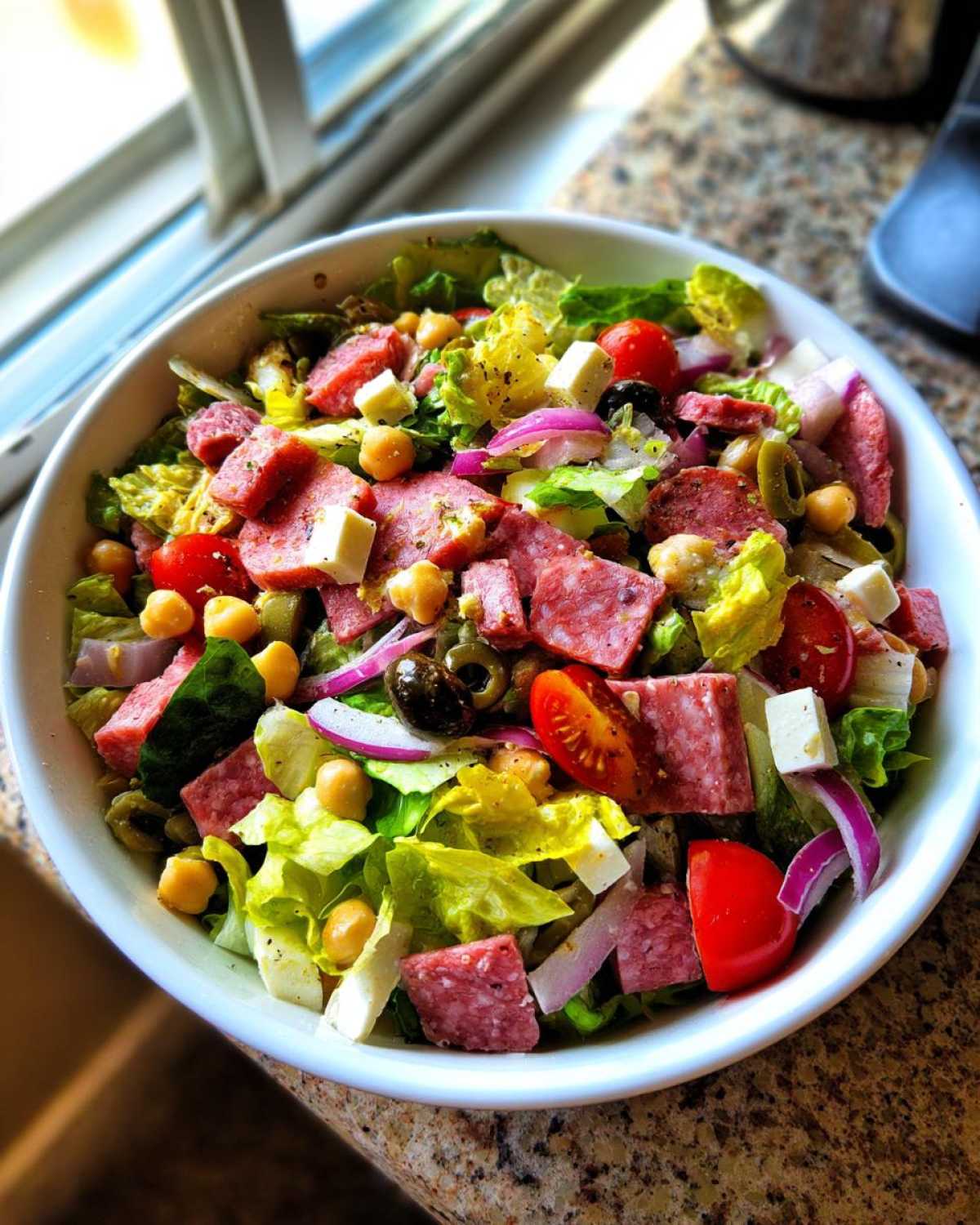 A close-up view of a colorful italian chopped salad in a white bowl, featuring salami, cheese, chickpeas, and vegetables.