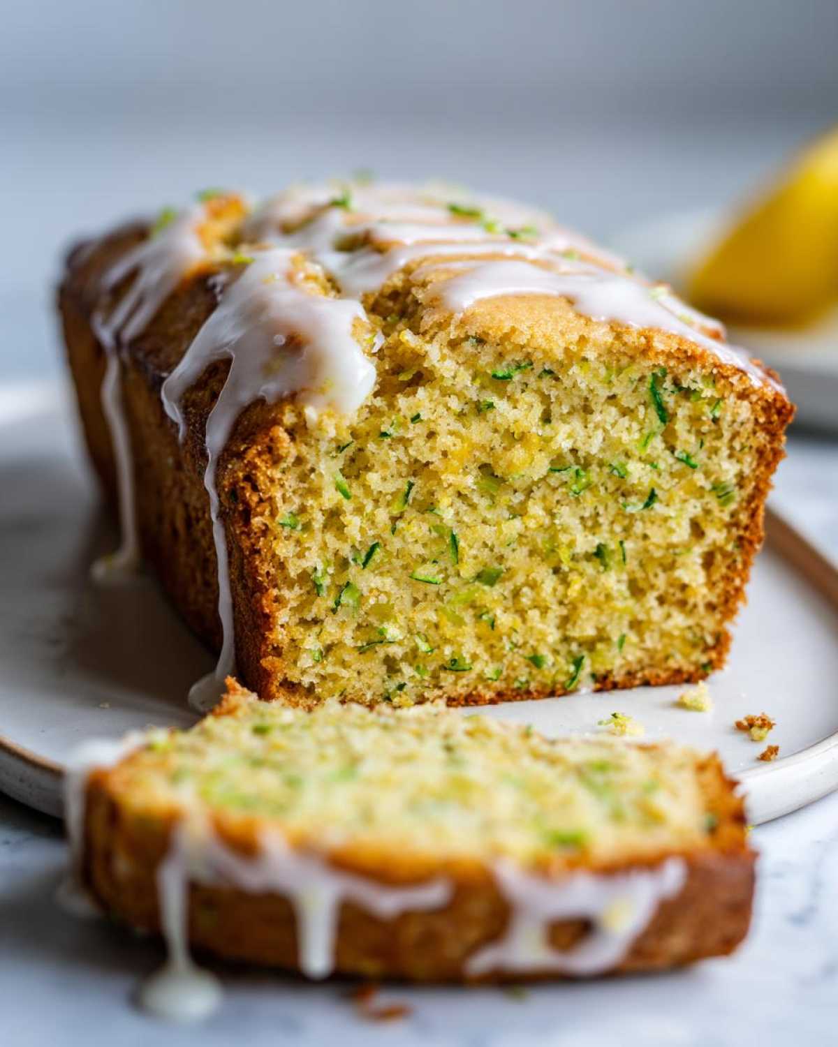 A whole loaf of lemon zucchini bread with a slice cut and placed in front, drizzled with white lemon glaze.