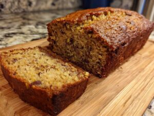A whole loaf of golden-brown zucchini banana bread with a single slice cut and placed in front of it on a wooden cutting board.