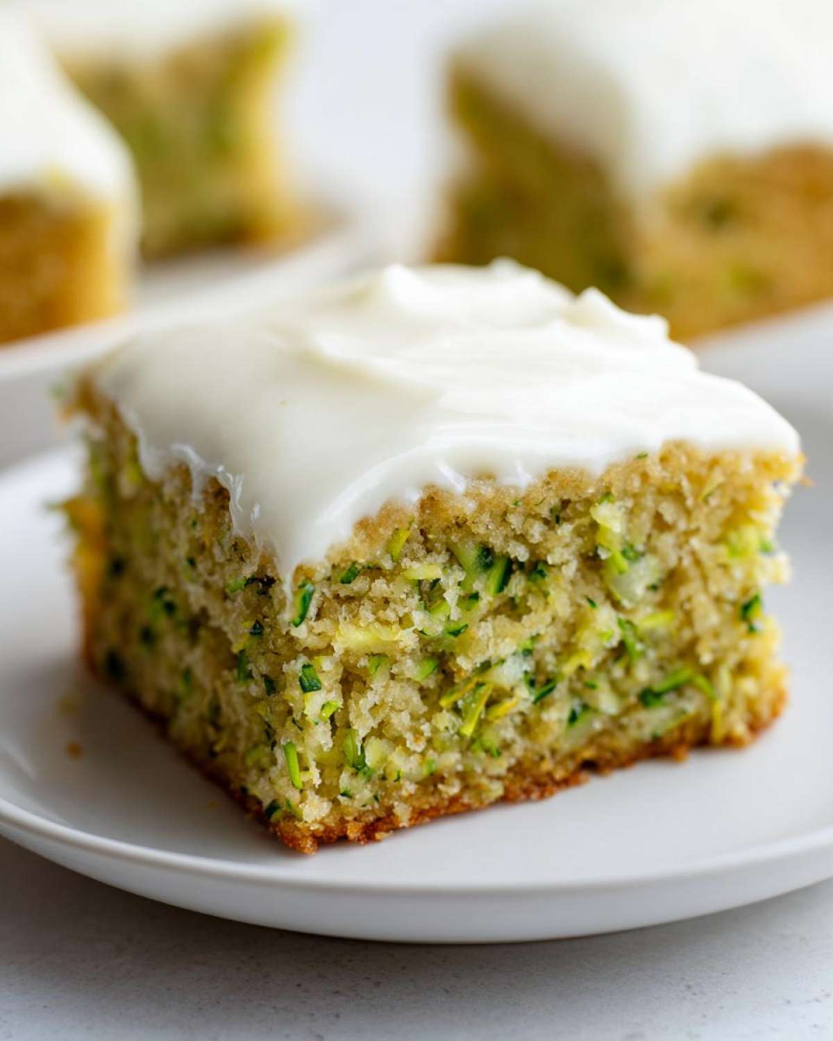 Close-up of a single square zucchini bar topped with thick white cream cheese frosting on a white plate.