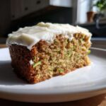 A close-up of a slice of moist zucchini cake with visible green zucchini pieces, topped with thick white cream cheese frosting.
