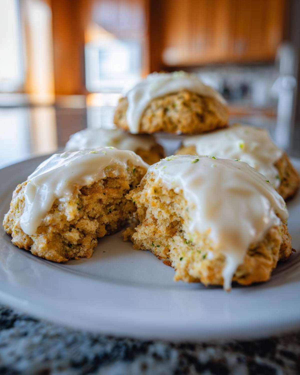 Close-up of several zucchini cookies on a white plate, topped with a creamy white glaze and lime zest. One cookie is broken in half.