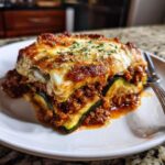 A close-up shot of a hearty slice of zucchini lasagna on a white plate, showing layers of zucchini, meat sauce, and melted cheese, garnished with herbs.