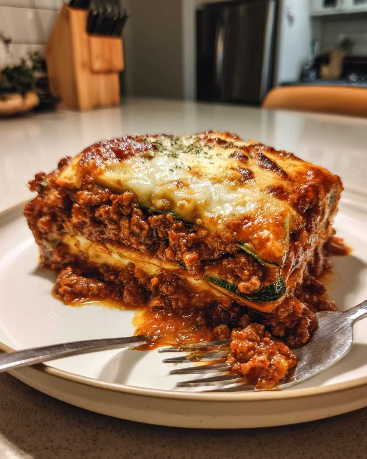 A close-up shot of a generous slice of zucchini lasagna on a white plate, showing layers of meat sauce, zucchini, and melted cheese.