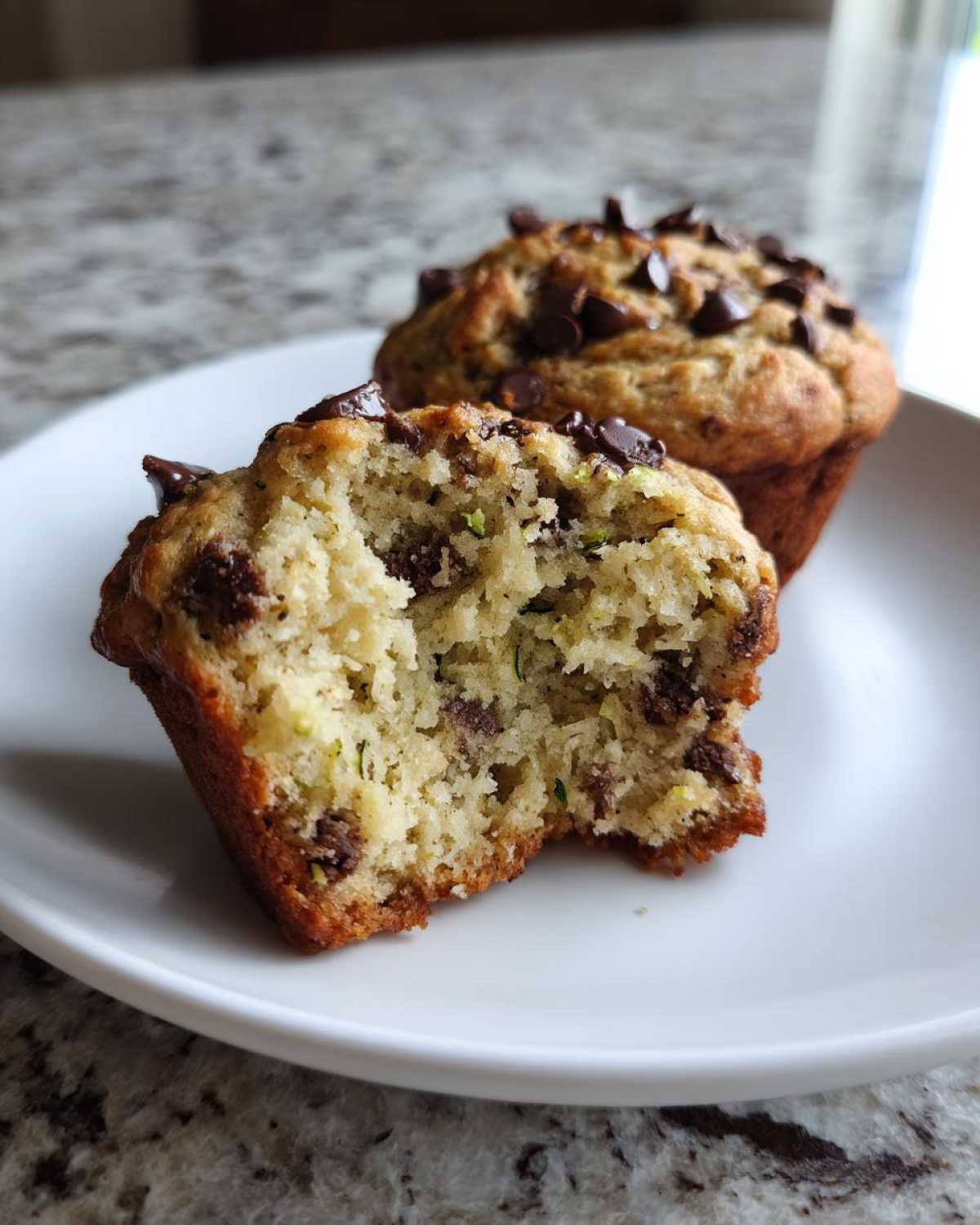 Close-up of a bitten zucchini muffin with visible chocolate chips and green zucchini flecks, on a white plate.