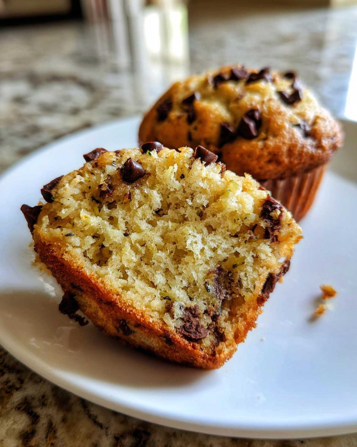 Close-up of a halved zucchini muffin on a white plate, showing its moist interior, flecks of zucchini, and chocolate chips. Another whole zucchini muffin is blurred in the background.