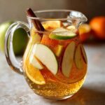 A glass pitcher filled with apple cider sangria, featuring apple and orange slices and a cinnamon stick.
