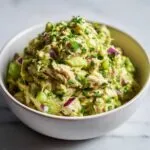 Close-up of a white bowl filled with creamy avocado tuna salad, garnished with fresh parsley and visible red onion.