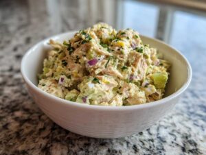 Close-up of a white bowl filled with creamy avocado tuna salad, garnished with fresh herbs and visible red onion.