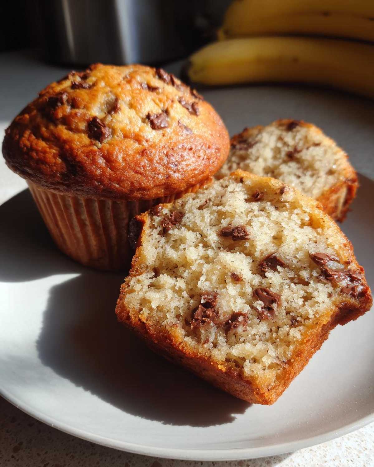 A whole banana protein muffin and a halved one on a plate, showing the fluffy texture and chocolate chips.