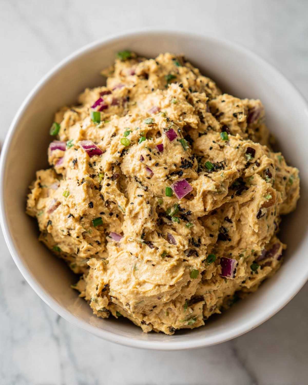 Close-up of a white bowl filled with creamy chickpea tuna salad, garnished with red onion, green onions, and black sesame seeds.
