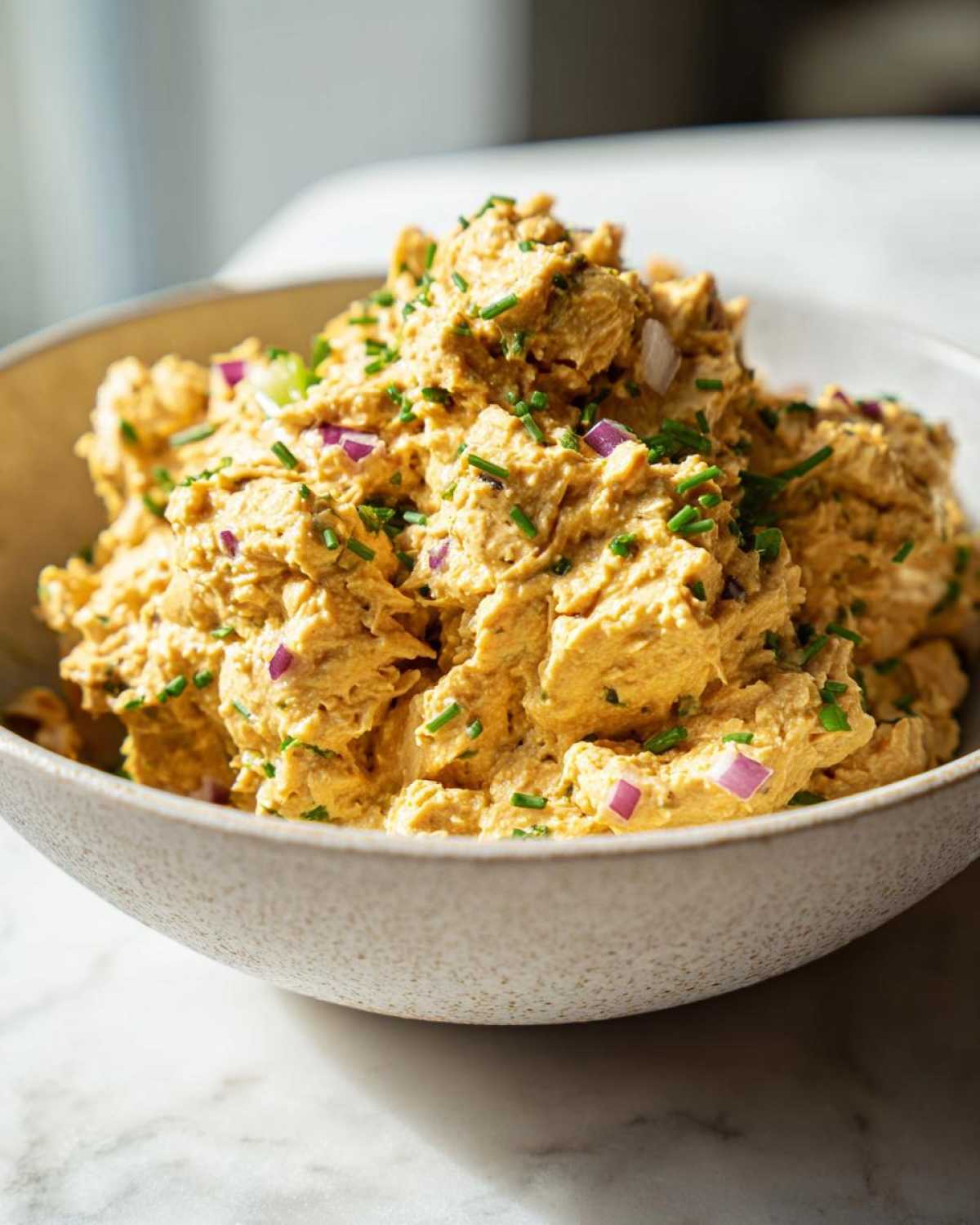 Close-up of a bowl filled with creamy chickpea tuna salad, garnished with chopped red onion and chives.