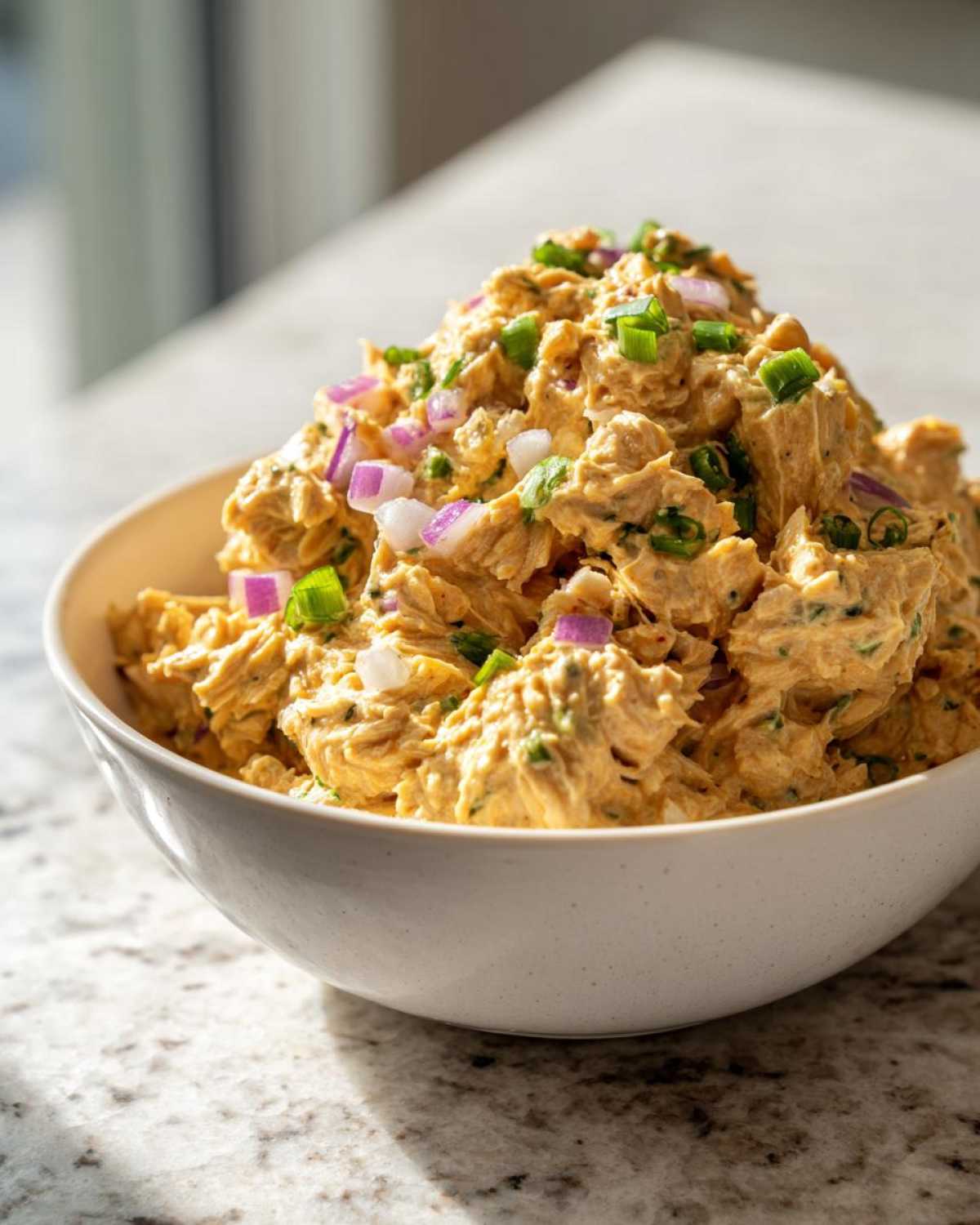 A close-up of a white bowl filled with creamy chickpea tuna salad, garnished with chopped red onion and green onions.
