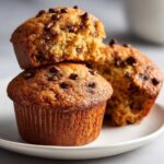 A stack of two golden-brown chocolate chip protein muffins on a white plate, with the top muffin broken open.