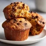 A stack of two golden-brown chocolate chip protein muffins on a white plate, with the top muffin broken open.