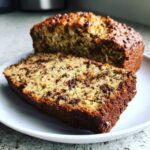 A slice of moist zucchini bread with chocolate chips, next to the full loaf, on a white plate.