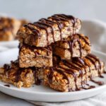 A close-up stack of several homemade protein bars, generously drizzled with dark chocolate, on a white plate.