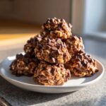 A close-up shot of a stack of chocolate peanut butter protein bites with visible oats and chocolate chips on a white plate.