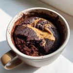 Close-up of a chocolate protein mug cake in a white mug, topped with a swirl of melted peanut butter.