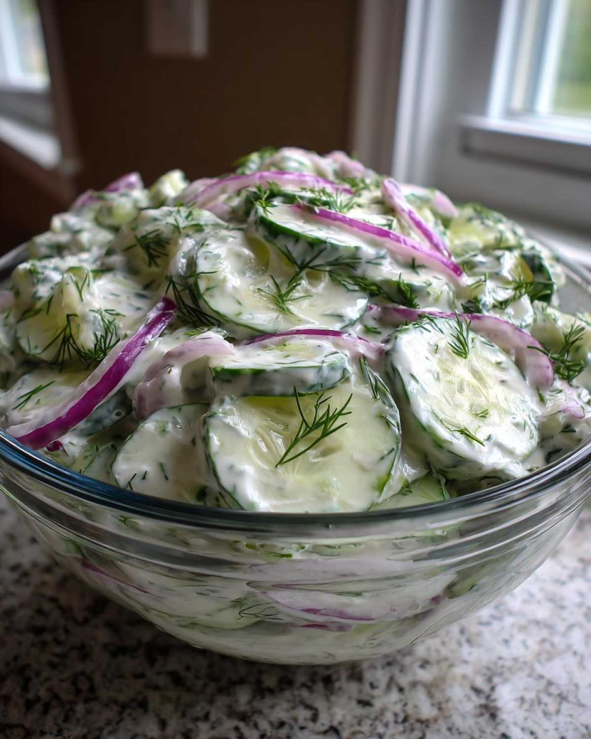 A large glass bowl filled with creamy cucumber salad, featuring sliced cucumbers, red onion, and fresh dill.