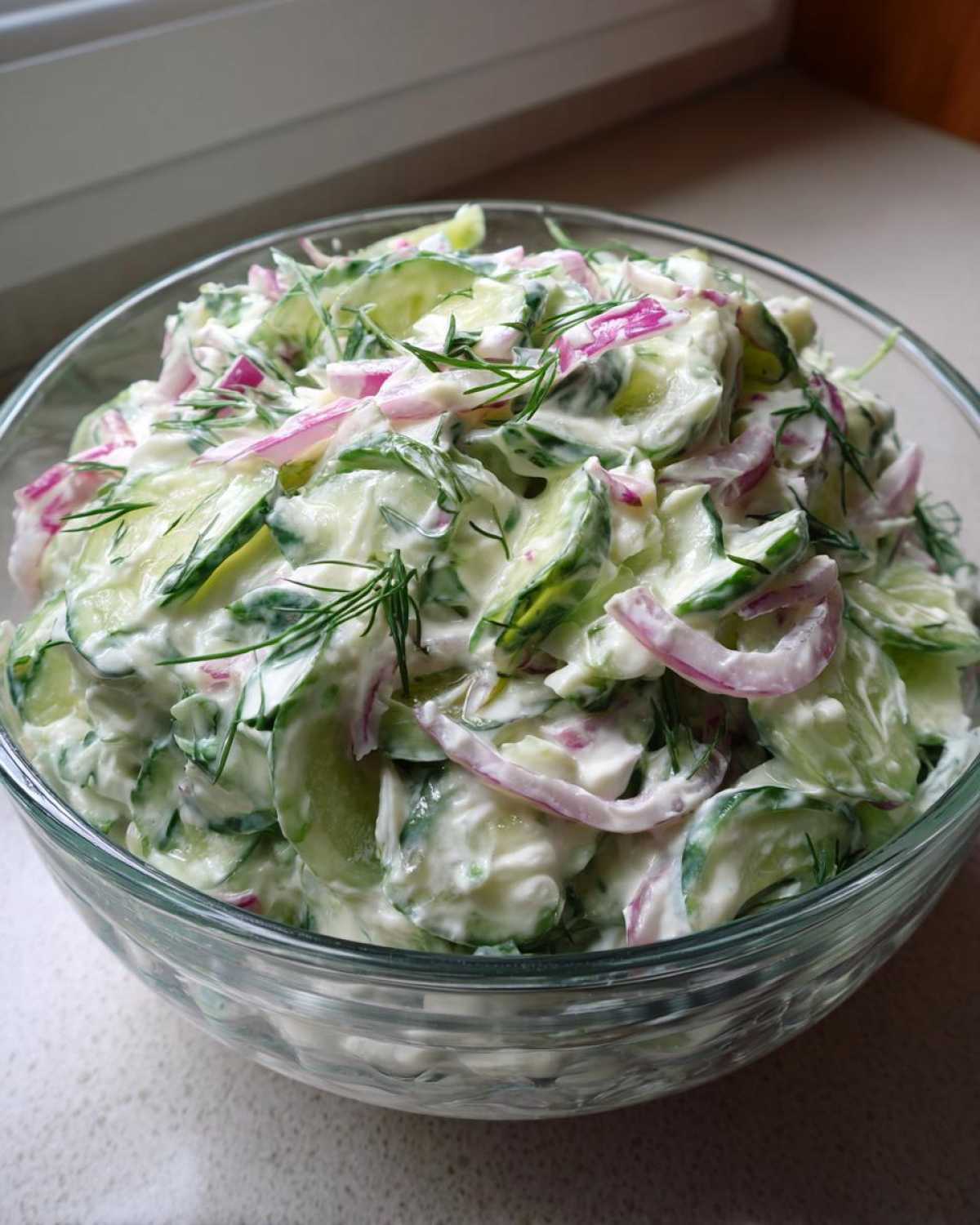 A clear glass bowl filled with creamy cucumber salad, featuring sliced cucumbers, red onions, and fresh dill.